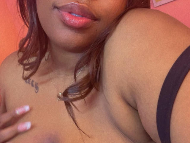 NatyLm with brown skin, pink glossy lips, and wavy hair, showing her bare shoulder and black strap, her hand on her tit, and a small necklace.