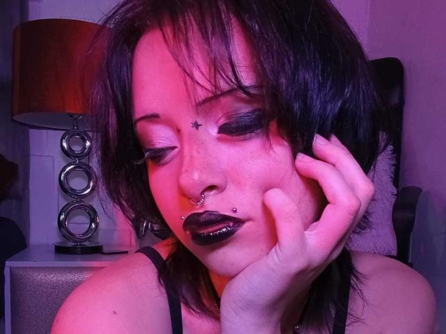 ViolettClay hat dunkles Haar, schwarzen Schminke, einen Stern auf der Stirn, Nasenringe, schwarzen Lippenstift und berührt ihr Gesicht. Dabei ist eine rosa und violette Beleuchtung zu sehen. Ihre Augen sind geschlossen..
