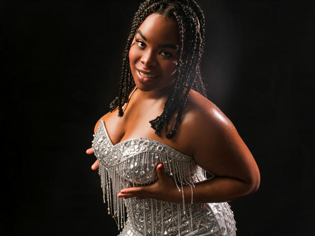 EbonyHenner, avec une peau foncée et brillante, souriante, portant une robe scintillante sans bretelles, touchant ses seins volumineux et brillants avec ses mains, cheveux tressés, fond sombre, éclairage de studio..