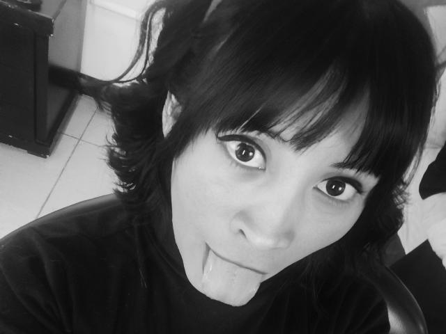 IssisDiosa with black hair, sticking out her tongue, and large eyes in a black-and-white selfie, wearing a dark shirt, looking up at the camera.