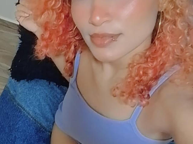 NaomyeLm, avec ses cheveux orange bouclés, portant un débardeur de sport violet, montrant ses seins, un petit piercing au nez, et la bouche légèrement ouverte avec une expression espiègle, assise sur un jean bleu..