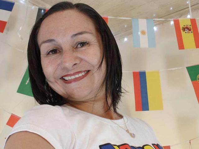 LatinLola, con i capelli neri e corti, sorride, indossa una camicia bianca, rossetto rosso e una collana, ha un viso carino, leggermente lentigginoso e un seno di medie dimensioni..