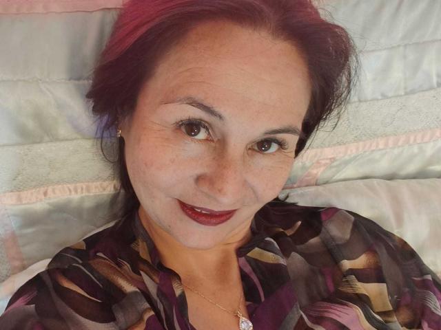 LatinLola tiene el cabello teñido de rosa, ojos marrones y lápiz labial rojo oscuro. Lleva una camisa estampada con colores púrpura, marrón y negro, y un pequeño collar dorado..