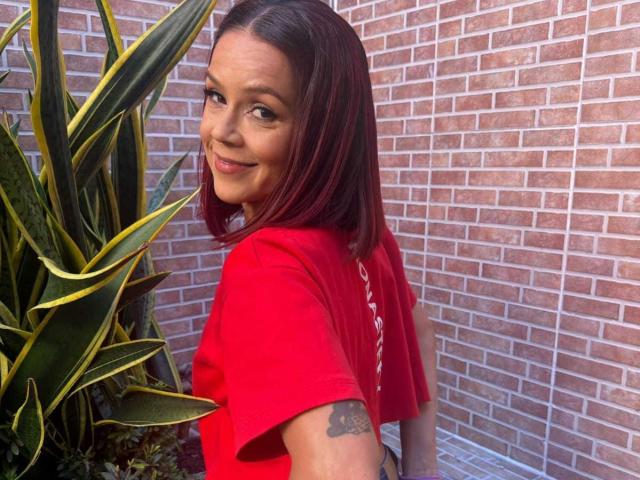 SharonBellarie, portant une chemise rouge, souriante, regardant en arrière, avec des yeux marron et des cheveux roux-brun, un petit tatouage sur le bras droit, debout près d'une plante serpent verte et jaune, avec un mur de briques en arrière-plan..