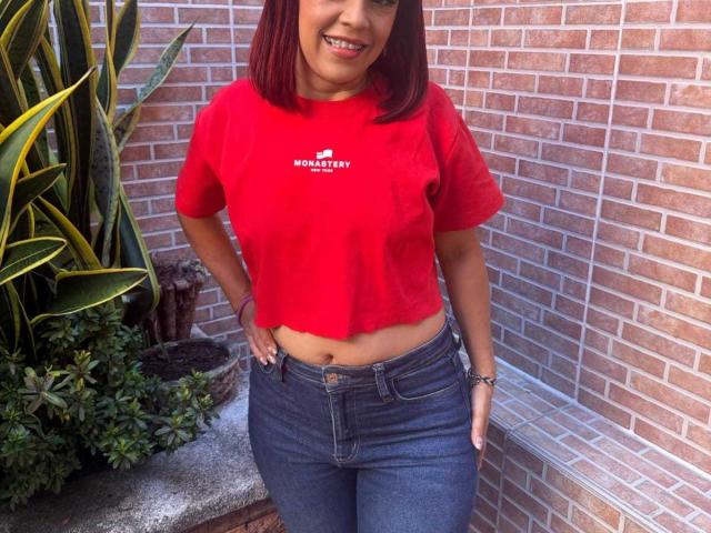 SharonBellarie with red shirt, showing her belly button, wearing blue jeans, hands on hips, smiling, medium brown skin, red hair, standing in front of brick wall.