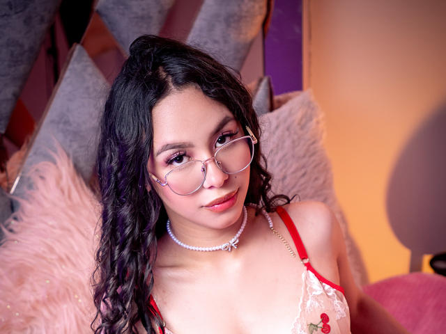Daphne Valucci, com cabelos pretos cacheados, óculos, um sutiã de renda vermelho e um colar de pérolas, olhando para a câmera com um sorriso discreto, mostrando seus seios. Iluminação suave e quente..