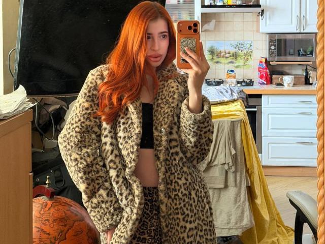 RosioRaschko, con i capelli rossi, indossa un cappotto di pelliccia a stampa leopardo, un top corto nero e pantaloni a stampa leopardo, e sta scattando un selfie allo specchio in una cucina. I seni di RosioRaschko sono parzialmente visibili..