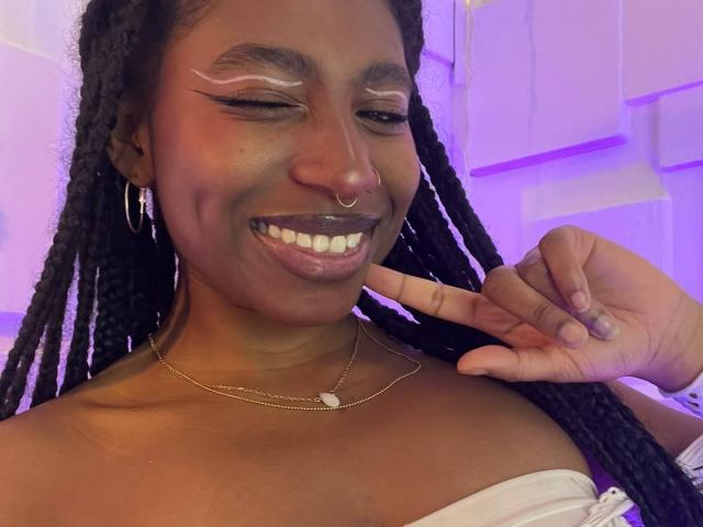 QueenPerlaa with dark skin, smiling with white eyeliner, nose ring, and gold necklace, winking, showing white teeth, and pointing to her face with her finger.