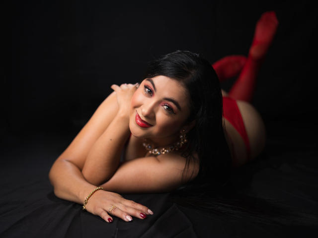 EmilyaBlaire, lying on her stomach, smiling, wearing red panties and red stockings, her ass up, black hair, red lipstick, gold bracelet, pearl necklace, black background.