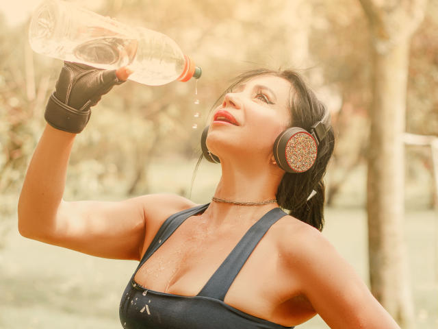HelenHard drinking from a bottle, wearing black gloves, headphones, and a choker, with sweat on her tits and a sexy smile, in a sunny outdoor setting.