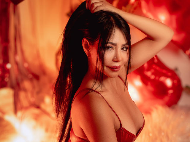 HelenHard with long black hair, red lingerie, and visible tits, holding her ponytail, smiling, in warm red lighting. Her ass is slightly visible in the background.