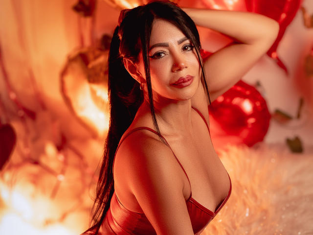 HelenHard with long black hair, red lingerie, and visible tits, posing with hand in hair, glowing red light, sexy smile, warm orange-red background.