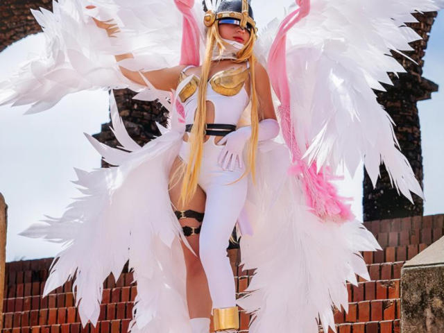 CyberZoe a de longs cheveux blonds, des ailes d'ange blanches, une armure dorée et un casque. Elle porte une combinaison blanche, des détails en plumes roses et des bottes dorées, et se tient à l'extérieur..