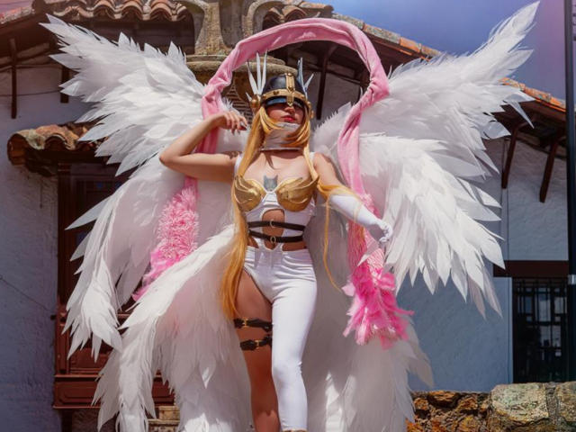 CyberZoe ha delle enormi ali bianche da angelo, piume rosa e bianche, lunghi capelli biondi, un pezzo dorato sul petto, una tuta bianca e un laccio sulla coscia, con un accessorio a forma di piuma rosa, in versione cosplay..