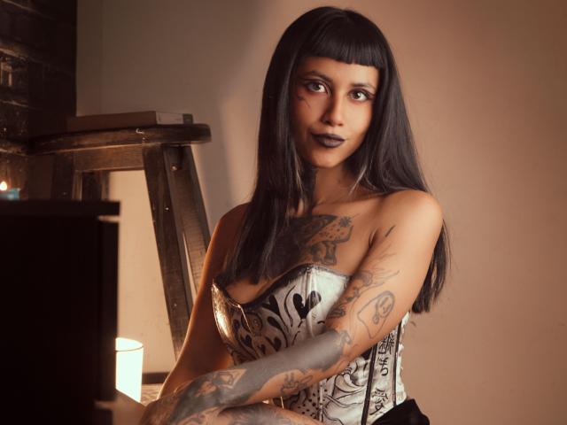 NyxVega with long black hair, dark lipstick, tattoos on arms and chest, wearing a white corset with black floral patterns, looking at camera, candle lit in background.