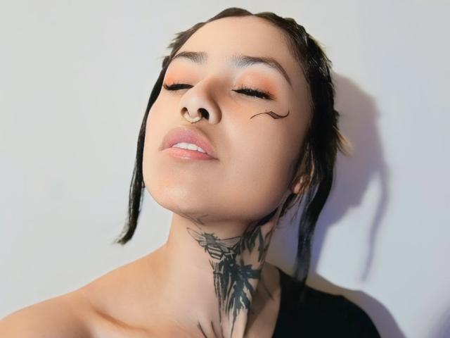 NyxVega with black braided hair, nose ring, eyeliner wings, closed eyes, subtle makeup, black tattoo on neck, light skin, shadow on white wall, relaxed expression.