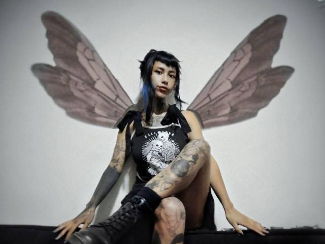 NyxVega with tattoos, black dress, and boots, sitting with legs crossed, wearing fairy wings, looking at camera, dark blue hair, pale skin, and a serious expression.