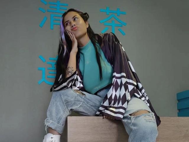 NyxVega, con el pelo largo y negro y un tatuaje en su brazo derecho, está sentada sobre una caja de madera, vistiendo una chaqueta de estampado blanco y negro, una blusa color turquesa y jeans rotos..