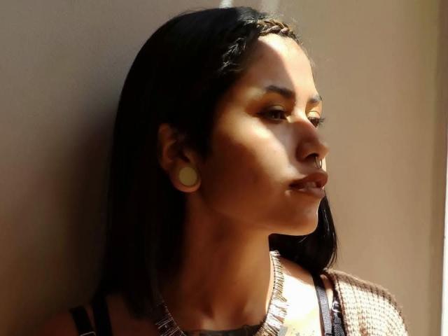NyxVega con capelli scuri, una treccia sulla testa, trucco giallo sugli occhi, grandi tappi per le orecchie, un maglione marrone, che guarda a destra, luce solare soffusa, una leggera ombra sul lato destro del viso..