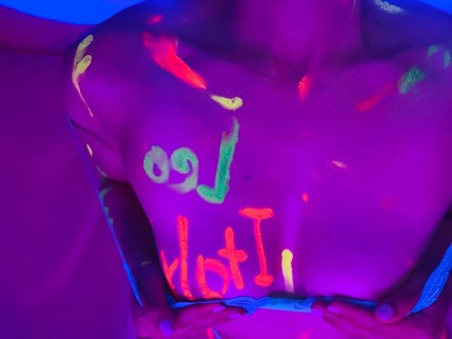 BelladonnaLips' glowing, neon-painted tits with colorful letters and symbols, hands below, in a purple-blue illuminated setting, highlighting her bare, chest-focused, illuminated body.