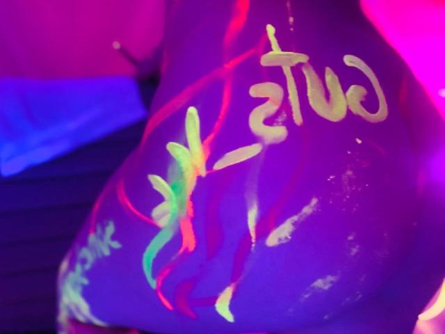 Glow-in-the-dark paint on BelladonnaLips' butt, featuring neon green, red, and yellow scribbles. Purple and pink neon lights in the background. Close-up, focusing on painted buttocks.