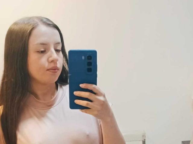 VioletaNova with long brown hair, light skin, and small tits, wearing a pink top, holding a blue phone, taking a selfie, looking down, neutral expression.
