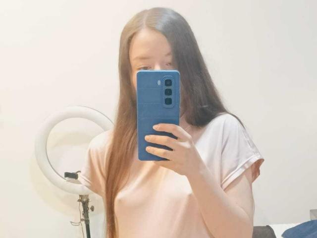 VioletaNova with long brown hair, pink t-shirt, holding blue phone, covering face, small tits, ring light in background, mirror selfie.