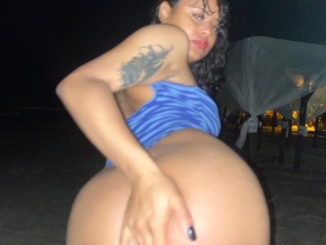 ChiaraBella with curly black hair, blue zebra-pattern bikini top, tattoo on right arm, holding her big ass, looking back at the camera, night, dark background.
