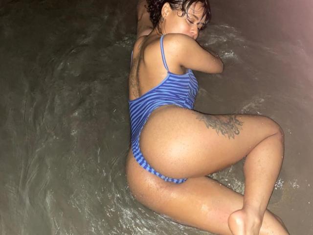 ChiaraBella is lying in the water, wearing a blue striped one-piece, with her ass up and her back to the camera, showing a tattoo on her right thigh.