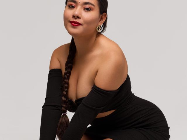 HannaGoez with big tits, dark braided hair, red lipstick, black off-shoulder dress, gold earrings, crouching, smiling, light skin, curvy, attractive.