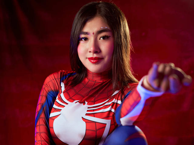 HannaGoez with medium tits, wearing a red Spider-Man costume, pointing with a blue-gloved hand, red lipstick, straight brown hair, and a subtle face decoration against a red background.