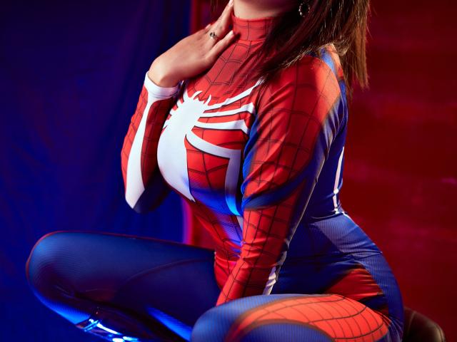HannaGoez in a Spider-Man costume, sitting, hand near neck, showing her tits, red and blue suit, white spider web, blue leggings, red and blue background, studio lighting.