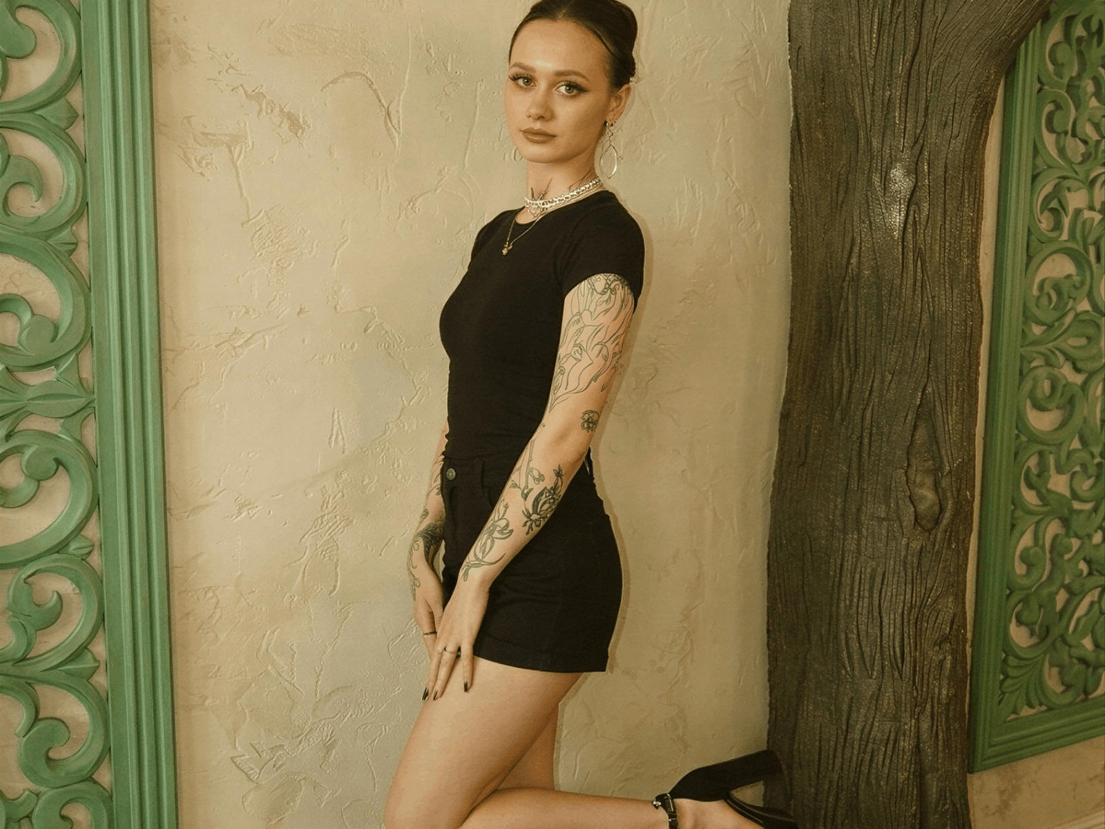 Krishnahabicht stands against a wall, wearing a tight black dress, showing tattoos on arms and legs, with a tree to the right, and green ornate frames on the sides.