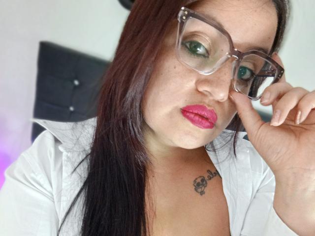 Ginebra Vargas mit dunkelbraunen Haaren, grünen Augen, pinkem Lippenstift, klaren Brillen, einem weißen Hemd und einem Tattoo auf der Brust, die ihre Brille mit ihrer rechten Hand justiert..