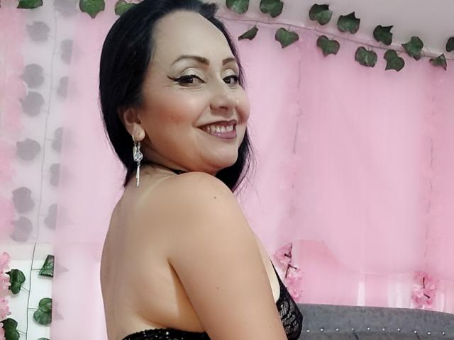 Zaria69, sonriendo con cabello negro, vistiendo un sujetador de encaje negro, mostrando su espalda y hombros desnudos, con aretes colgantes, en una habitación de color rosa con hojas verdes..