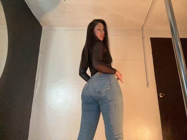 EmmaLombardi with long black hair, wearing a sheer black top and tight blue jeans, showing off her big ass and curvy butt, standing in a room.