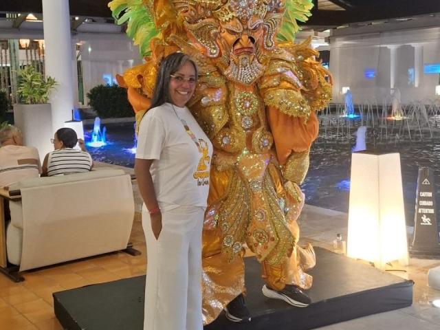 Laila Castillo está de pie junto a una enorme máscara china de dragón, brillante, naranja y dorada, con plumas verdes, vestida con pantalones blancos y una camisa blanca, sonriendo a la cámara..