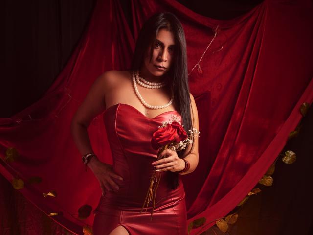 AfroditaDoir with long black hair, wearing a red leather strapless dress, pearl necklace, holding red roses, standing with hand on hip, dark red draped background, sexy, confident.