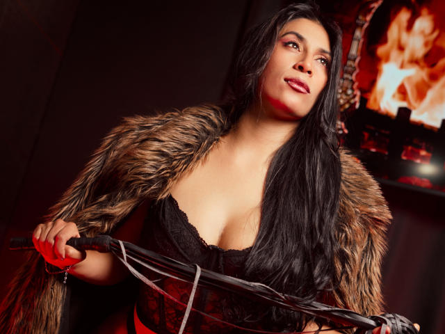 AfroditaDoir with long black hair, wearing a fur coat and black lace corset, holding a whip, looking up with a slight smile, red light in the background.