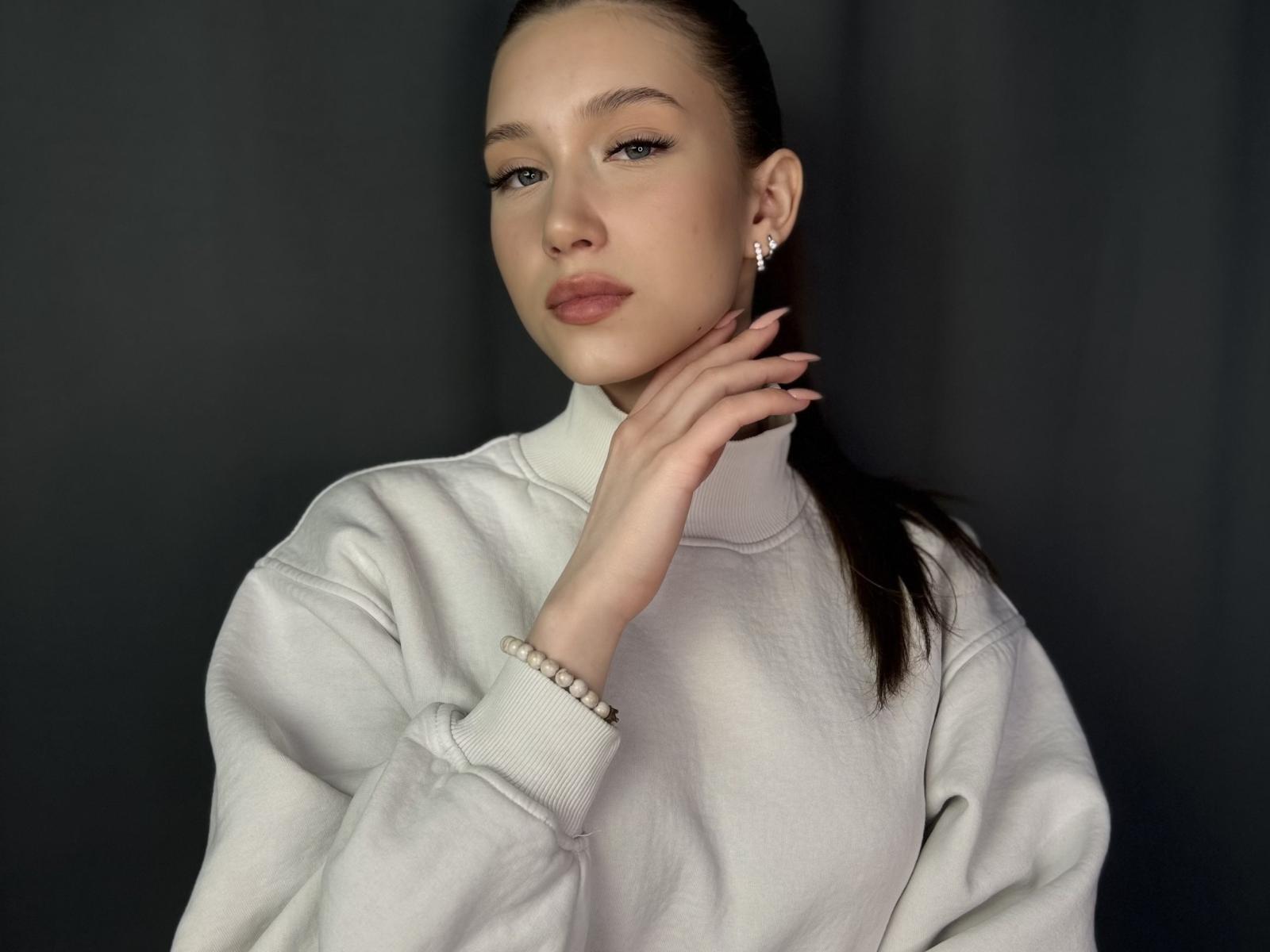 JanethNgov, avec une peau claire, des cheveux bruns attachés en queue de cheval, portant un pull blanc à col roulé, se tenant le cou, regardant la caméra, fond gris, bracelet au poignet gauche, boucles d'oreilles..