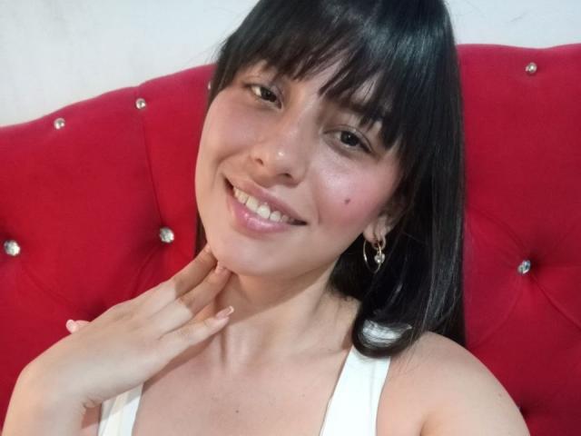 MadissonFontaine with black hair and bangs, smiling, white tank top, hand on neck, red tufted headboard with sparkles, pink lips, brown eyes, small mole on left cheek.