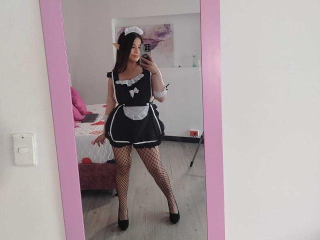 Melani Becker, vêtue d'un uniforme de femme de ménage noir et blanc, avec des bas résille et des talons noirs, se tient devant un miroir, montrant ses fesses et ses seins, tout en tenant un téléphone..