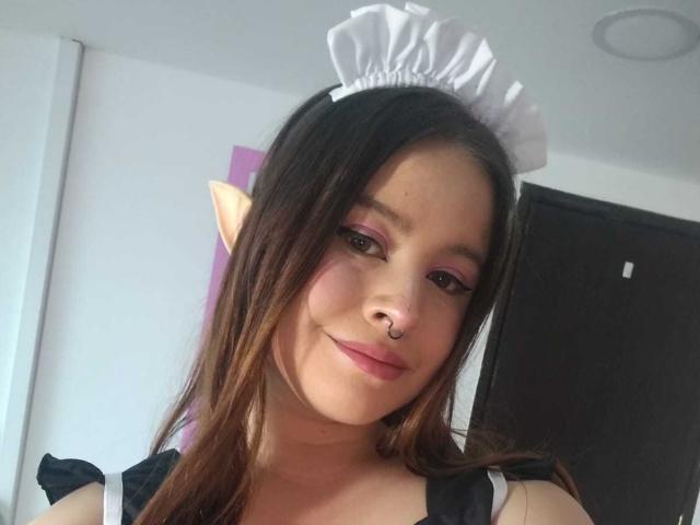 Melani Becker, con orecchie a punta da elfo, indossa una cuffia da cameriera nera e bianca, trucco rosa per gli occhi, un anello al naso e un abito nero, sorridendo dolcemente alla fotocamera..