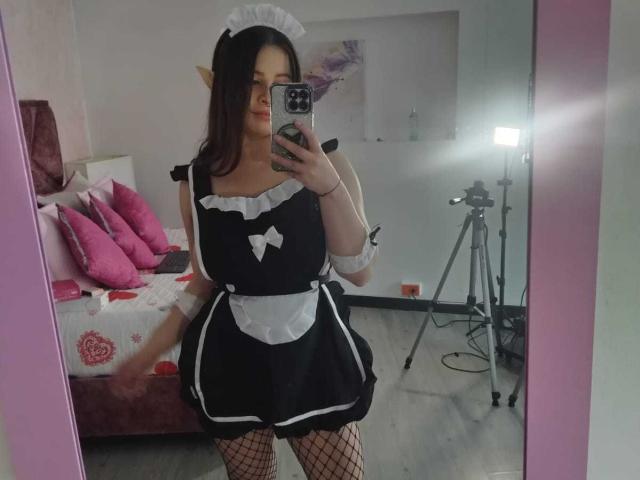 Melani Becker, con orecchie a punta, indossa un abito da cameriera nero e bianco, calze a rete e un copricapo bianco, e sta scattando un selfie allo specchio, mostrando i suoi seni e il suo piccolo sedere..
