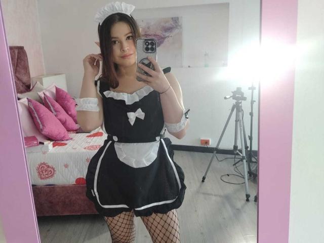 Melani Becker, in un abito da cameriera bianco e nero con calze a rete, tiene un telefono e si trova di fronte a uno specchio. Ha i capelli castani, un viso carino e piccoli seni..