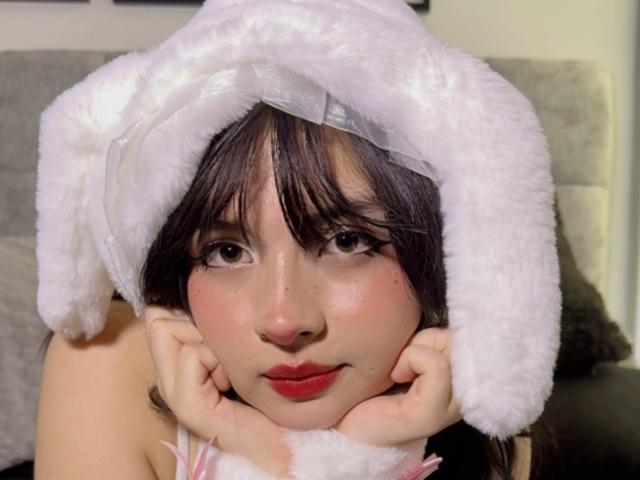 LiliithD, avec un chapeau rose et duveteux, les mains sur les joues, les yeux marron, les lèvres rouges et les cheveux brun foncé, a l'air mignonne et légèrement rougie. Ses seins ne sont pas visibles..