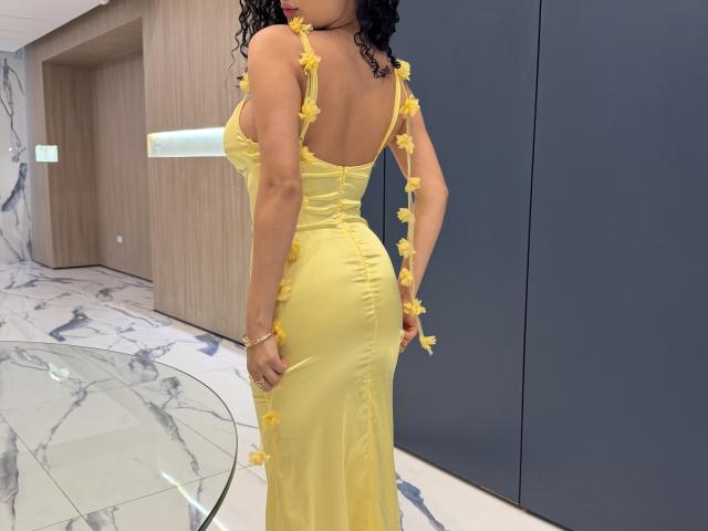 Aurora Ferretti, usando um vestido amarelo com detalhes de flores amarelas, está de costas para a câmera, mostrando suas nádegas, cabelos pretos cacheados e uma pulseira amarela..