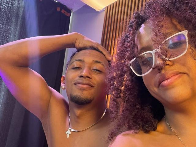 LilithAndAsmodeo, com cabelo cacheado, óculos e sem camisa, está ao lado de LilithAndAsmodeo, um homem sem camisa, com um colar, a mão na cabeça e sorrindo. Ambos têm pele morena..