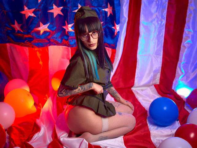 TatsumiLee with glasses, black hair, and blue highlights, wearing a military cap and green shirt, kneeling, showing her tattooed arm and white fishnet stockings, with colorful balloons.