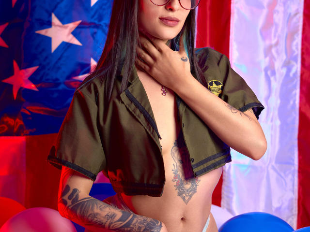 TatsumiLee with tattoos, green crop jacket, glasses, and bare tits, hand on neck, American flag background, colorful balloons, tattoos on chest and arm, long black hair, subtle smile.